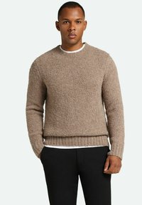 Brązowy sweter dzianinowy o fakturowanej powierzchni, okrągłym dekolcie i ściągaczu na dole. Nosi się go na białej koszuli, w połączeniu z czarnymi spodniami.