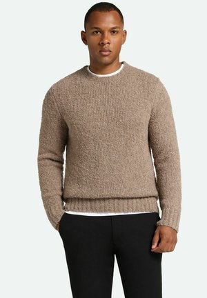 Maglione brown in maglia con superficie texturizzata, collo rotondo e orlo a coste. Indossato sopra una camicia bianca, abbinato a pantaloni neri.