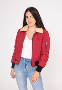 Veste bomber rouge avec un col en fourrure beige, poches zippées, poignets et taille côtelés. Portée sur un haut blanc et associée à un jean bleu.