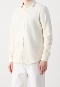 Camicia button-up color crema chiaro realizzata in tessuto texturizzato, con colletto a punta e ricamo del logo discreto. Maniche lunghe e orlo curvo.