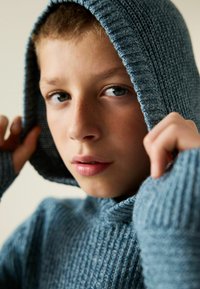 Strick-Hoodie in sanftem Blau, mit einem strukturierten Rippenmuster und einer großen Kapuze, die teilweise das Gesicht bedeckt, während die Hände den Stoff greifen.