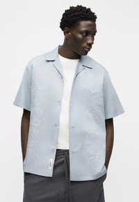 Άνδρας με ανοιχτό μπλε πουκάμισο με κοντά μανίκια και λευκή κεντητή σπείρα, λευκό εσώρουχο και σκούρα γκρι παντελόνια με κορδόνι, κοιτάζει δεξιά.