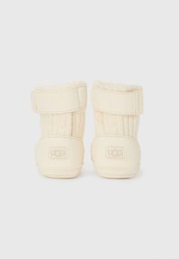 Bottes pour nourrissons couleur crème avec un dessus en tricot texturé doux, fermeture à sangle sécurisée et logo UGG visible à l'arrière.