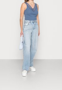 Blå linne med spetsdetaljer, högmidjade ljusblå jeans, vita plattformssneakers och en ljusblå väska i ena handen.