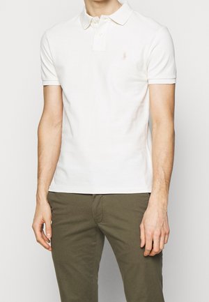 Homme portant un polo blanc ajusté à manches courtes et un pantalon vert olive, debout devant un fond clair uni.