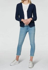 Blazer in maglia blu navy con due bottoni, abbinato a un top bianco con paillettes, jeans ankle azzurri chiari con dettagli in perle e sneaker bianche.