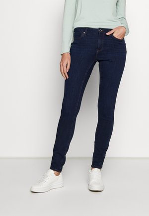 Vrouw draagt donkerblauwe skinny jeans, een lichtgroen shirt met lange mouwen en witte sneakers, staand met één hand in de zak.