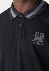 Zwarte polo met een gestructureerde stof, voorzien van een grijs gestreepte kraag, twee knopen en een textuur logo ontwerp op de borst.