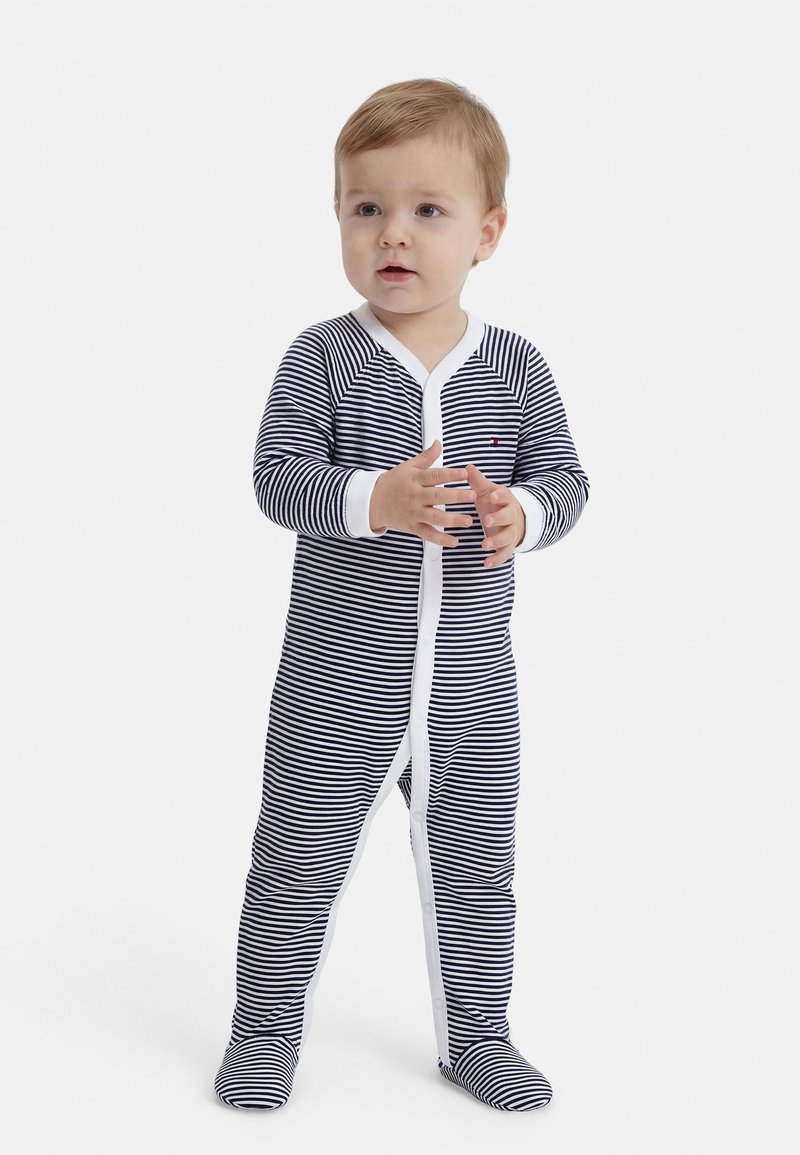 Μικρό παιδί που στέκεται, φορώντας μαύρη και λευκή ριγέ ολόσωμη πιτζάμα με λευκή επένδυση και κουμπιά μπροστά.
