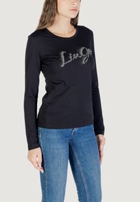 Un modello indossa una maglietta nera a maniche lunghe con un logo luccicante "LiveGo", abbinata a jeans blu, su uno sfondo neutro.