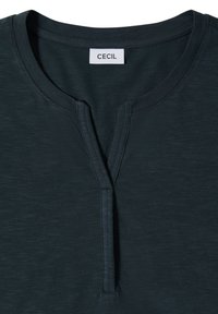 T-shirt vert foncé à col rond avec une fente courte sur le devant et des coutures visibles, avec une étiquette blanche portant le texte « CECIL » à l’intérieur du col.