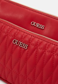 Neceser de cuero rojo con un patrón acolchado y el logo en tono plateado de "GUESS". Presenta un cierre de cremallera y una textura suave.
