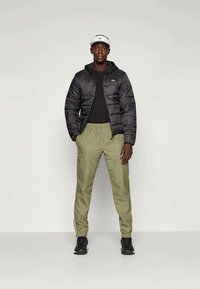 Casaco puffer preto com capuz, camisa preta de mangas longas, calças elásticas de verde claro e ténis pretos. Viseira branca na cabeça.