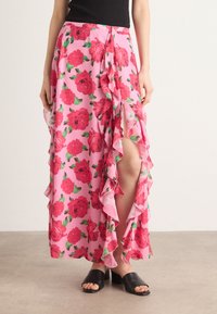 Jupe maxi florale rose avec des volants sur le côté et l'ourlet, ornée de grandes roses rouges et de feuilles vertes, fabriquée en tissu léger.