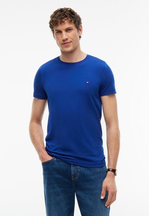 Junger Mann mit kurzen lockigen Haaren, der ein schlichtes blaues T-Shirt und blaue Jeans trägt, mit einer Hand in der Tasche und einer Uhr am Handgelenk.
