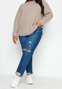 Kvinna i en överdimensionerad beige stickad tröja, slitna blå jeans med fransiga fållar och vita sneakers, som står framför en enfärgad vit bakgrund.