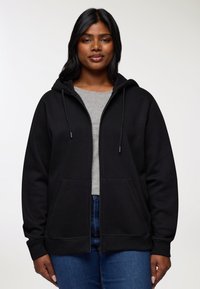 Kobieta nosząca czarną bluzę z kapturem zapinaną na zamek, pod którą ma szary T-shirt oraz niebieskie dżinsy, stojąca na tle jasnej, jednolitej ściany.