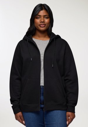 Donna con una felpa nera con zip sopra una maglietta grigia e jeans blu, in piedi contro uno sfondo chiaro e uniforme.