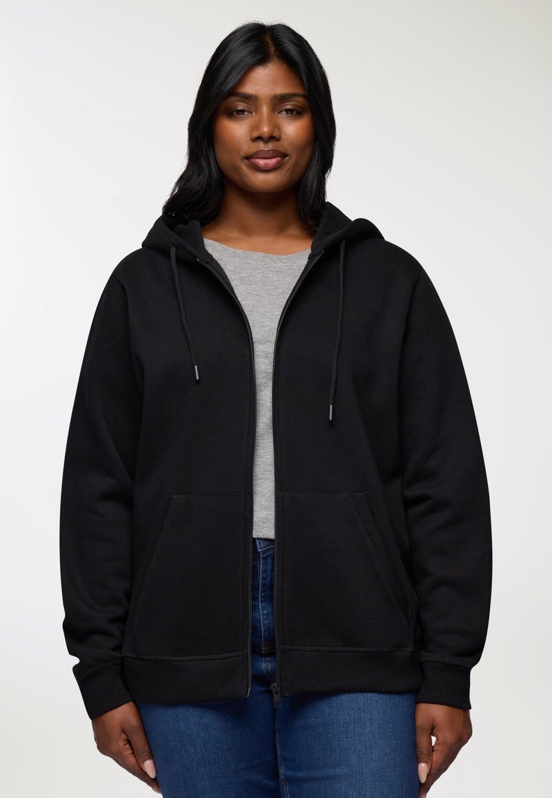 Kobieta nosząca czarną bluzę z kapturem zapinaną na zamek, pod którą ma szary T-shirt oraz niebieskie dżinsy, stojąca na tle jasnej, jednolitej ściany.