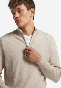 Jasno-beżowy sweter na zamek wykonany z teksturowanej dzianiny, z wysokim kołnierzem i ściągaczami na rękawach, z białą koszulą włożoną pod spód.