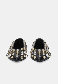 Zapatos planos negros con una forma alargada, adornados con tachuelas metálicas doradas y plateadas en toda la superficie. Textura suave y lados abiertos.