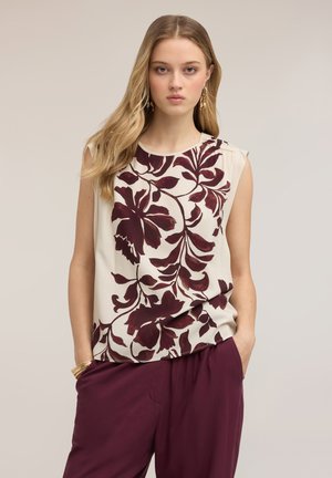 Jeune femme portant un haut crème sans manches avec un grand imprimé floral rouge foncé et un pantalon rouge foncé assorti, les mains dans les poches.