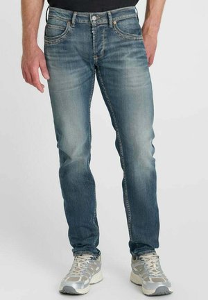 Uomo con jeans blu sbiaditi e scarpe da ginnastica grigie e bianche in piedi davanti a uno sfondo semplice.
