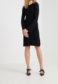 Femme portant une robe côtelée noire ajustée à manches longues, debout les mains jointes, avec des talons noirs pointus à brides de cheville.