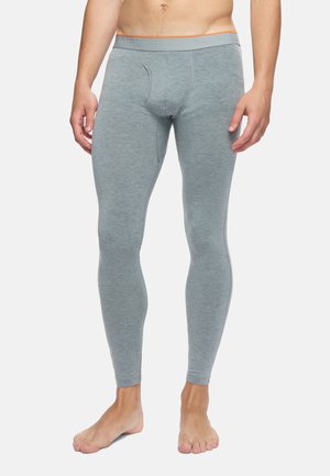 Homme portant un pantalon long de sous-vêtement ajusté gris clair avec une ceinture marquée Merrell, debout pieds nus sur un fond uni.