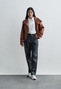 Kvinne iført en brun skinnjakke, hvit skjorte med knapper, svarte skinnbukser og hvite sneakers, stående mot en enkel lys bakgrunn.