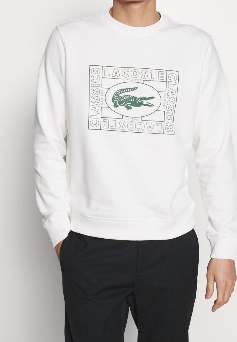 Homme portant un sweat-shirt blanc avec le logo crocodile vert Lacoste et les mots "Lacoste Classics Since 1927" dans un design encadré à l'avant.