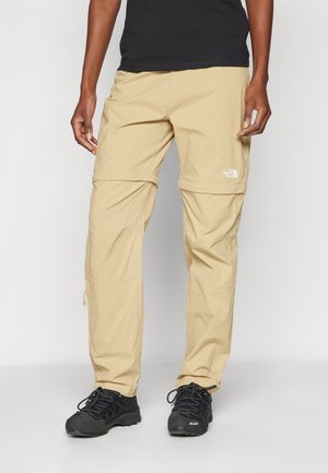 Beige kevyet zip-off cargo-housut, joissa on useita taskuja, joustava vyötärö ja merkkilogo alaleuassa; yhdistetty mustiin kenkiin.