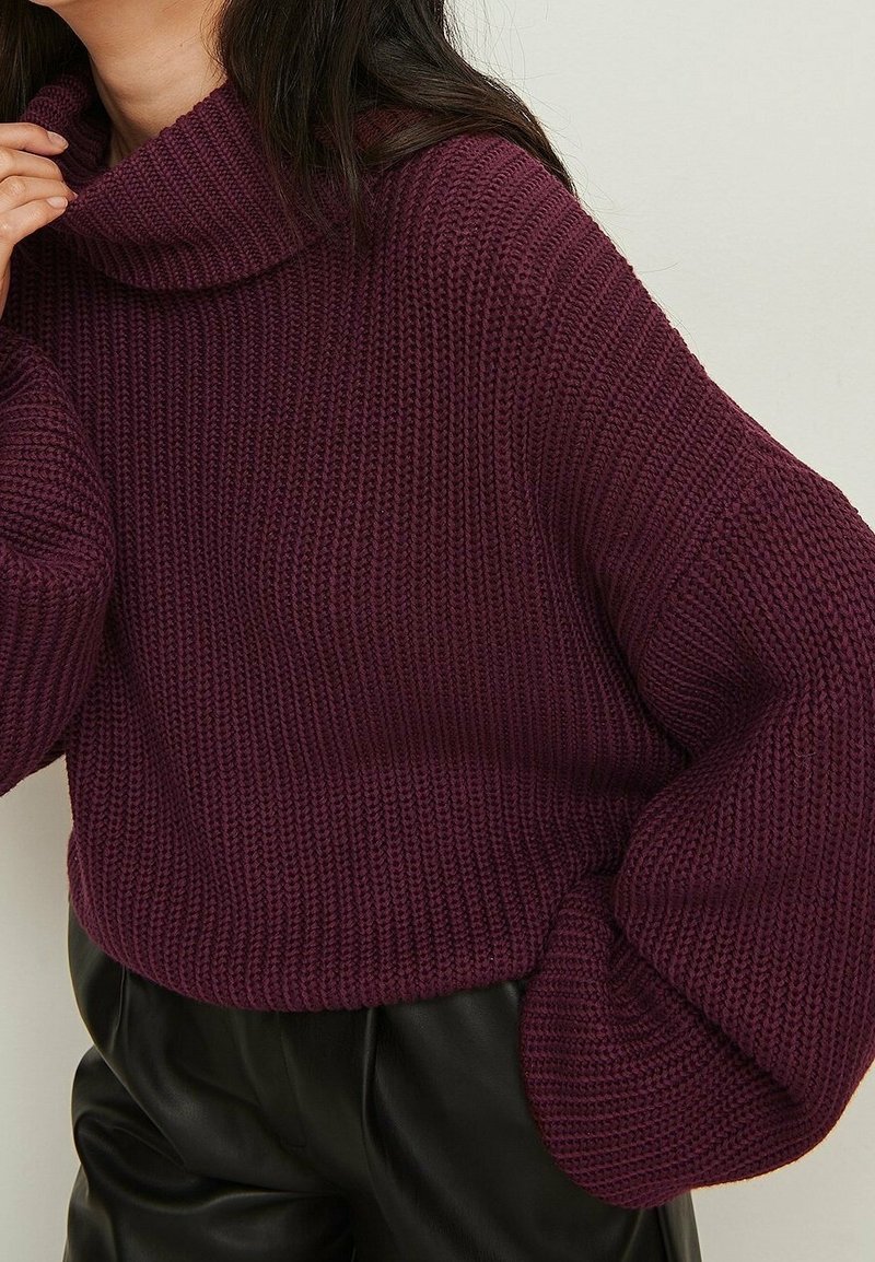 Femme portant un pull à col roulé bordeaux en maille épaisse avec des manches ballon et un pantalon en cuir noir sur un fond neutre.