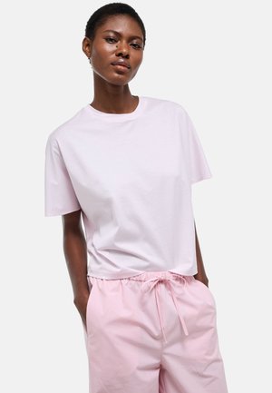 Femme à la peau foncée portant un t-shirt rose clair à manches courtes et un pantalon rose assorti avec un cordon, debout les mains dans les poches.