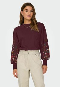 Felpa bordeaux con ricami floreali sulle maniche over, abbinata a pantaloni beige chiaro con vita elastica e tasche.