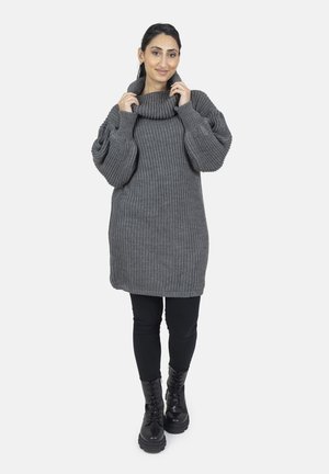 Kvinde iført en grå oversized strikket sweaterkjole, sorte leggings og sorte snørestøvler, der holder sweaterens krave med begge hænder.