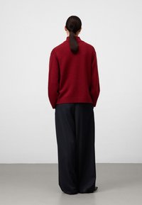 Vrouw met een donker paardenstaart draagt een rode gebreide trui en losse zwarte broek, staand met haar rug naar de camera tegen een effen achtergrond.