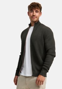 Mørkegrøn, struktureret zip-sweater med høj krave, der har en kontrasterende lynlås, båret over en hvid T-shirt og parret med lyse bukser.