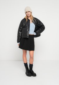 Νεαρή γυναίκα που φοράει μαύρο καπιτονέ μπουφάν, ανοιχτό μπλε κοντό τοπ, μαύρη φούστα, μπεζ πλεκτό καπέλο και μαύρα αρβυλάκια με κορδόνια και σκούρες κάλτσες.