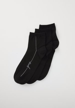 Chaussettes noires en tissu doux, avec un motif de lignes blanches et un logo en écriture cursive. Trois paires regroupées.