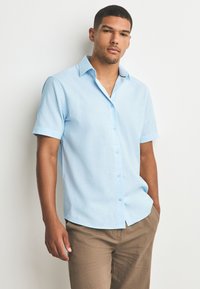 Mann mit kurzen Haaren, der ein hellblaues, kurzärmeliges Hemd mit Knöpfen und braune Hose trägt, steht vor weißem Hintergrund mit einer Hand in der Hosentasche.