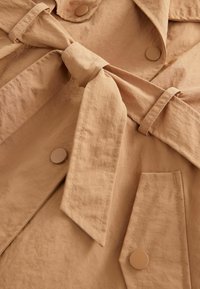 Gros plan d’un trench beige montrant une ceinture en tissu nouée, des boutons, un col et une poche avec rabat boutonné.