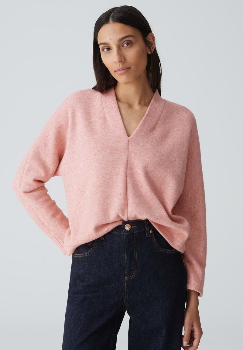 Helles rosa V-Ausschnitt-Pullover aus weichem Strickstoff, mit langen Ärmeln, lockerer Passform und kombiniert mit dunklen Jeans.