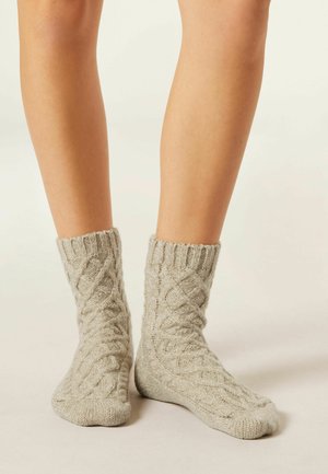 Chaussettes tricotées en gris clair avec un motif en câble texturé. Bord-côte en haut et zone de orteil lisse contrastée. Matière douce en mélange de laine.