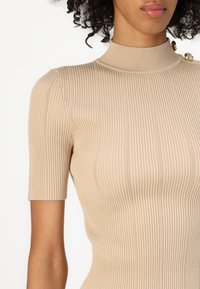 Beige ribbestrikket topp med korte ermer, høy krage og dekorative knapper på skulderen, med en tettsittende silhuett og teksturedetaljer.