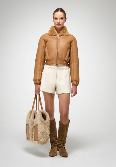 Giubbotto bomber in pelle color cammello con colletto in pelliccia, pantaloni corti crema, stivali marrone al ginocchio e una borsa morbida con dettagli in pelle.