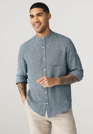 Button-up shirt in blue and white houndstooth pattern, featuring a banded collar, chest pocket, and rolled sleeves. Made of textured fabric.