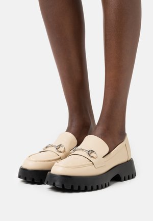 Mocassins en cuir beige avec un bout arrondi, accent en métal argenté, et une semelle épaisse en caoutchouc noir présentant des motifs de stries en vagues.