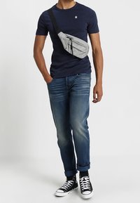 Szara nerka z zamkniętą kieszenią, noszona przez ramię; granatowy t-shirt, niebieskie dżinsy i czarne sneakersy wysokie z białymi podeszwami.