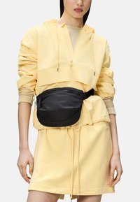 Borsa in nylon nera con una superficie liscia, caratterizzata da un design curvo e una tasca frontale con zip, indossata su una tuta leggera gialla.